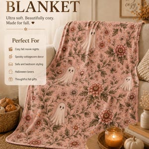 May include: A velveteen plush blanket featuring a repeating pattern of white ghosts, pink flowers, and green foliage on a light pink background. The text on the blanket includes Velveteen Plush BLANKET, Ultra soft. Beautifully cozy. Made for fall.