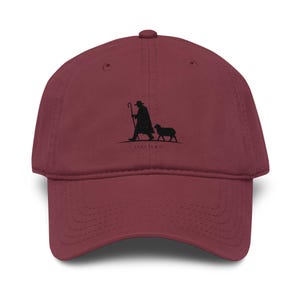 Könnte beinhalten: Eine kastanienbraune Baseballkappe mit einer schwarzen Stickerei, die einen Hirten mit einem Stab zeigt, der neben einem Schaf geht. Unter der Grafik steht der Text LUKE 15:4-7. Die Kappe hat einen gebogenen Schirm und eine strukturierte Krone.