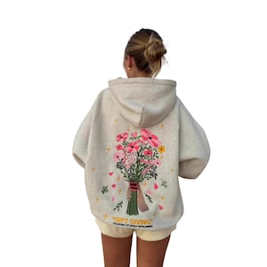 Può includere: Una felpa con cappuccio color avena caratterizzata da una grafica ricamata sul retro. Il disegno raffigura una mano che tiene un colorato bouquet di fiori rosa, rossi e gialli con steli verdi, circondato da piccole stelle gialle e dal testo GIFT GIVING.