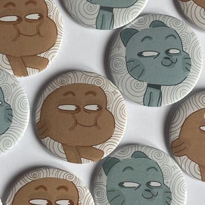 May include: A selection of circular pin badges with cartoon character designs. Some badges show a light brown character with a smiling face, while others feature a blue-grey cat-like character. Each badge has a white background with a spiral pattern.