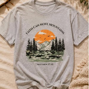 May include: A light grey t-shirt with a landscape design featuring mountains, trees, and a setting sun. The text "Faith Can Move Mountains" is arched above the design, with "Matthew 17:20" below.