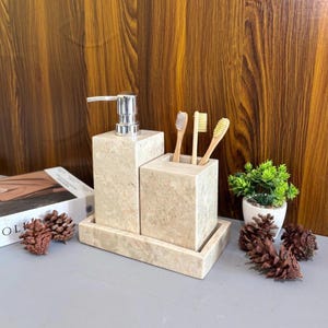 Può includere: Un set da bagno in marmo beige con un dosatore di sapone, un portaspazzolino e un vassoio. Il dosatore di sapone ha una pompa argentata. Tre spazzolini da denti in bambù sono nel supporto.