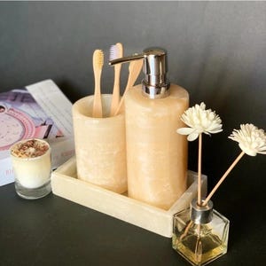 Può includere: Un set da bagno in pietra beige chiaro con dispenser di sapone, portaspazzolini e vassoio. Il set include spazzolini da denti in legno ed è completato da una piccola candela e un diffusore a bastoncini con fiori bianchi.