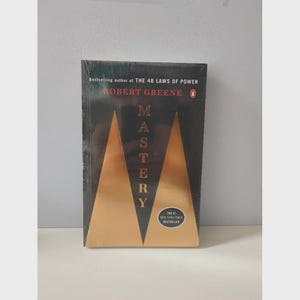 May include: A hardcover book titled "Mastery" by Robert Greene. The cover is black with two gold geometric shapes and the title in gold lettering. The text "The #1 New York Times Bestseller" is also visible.