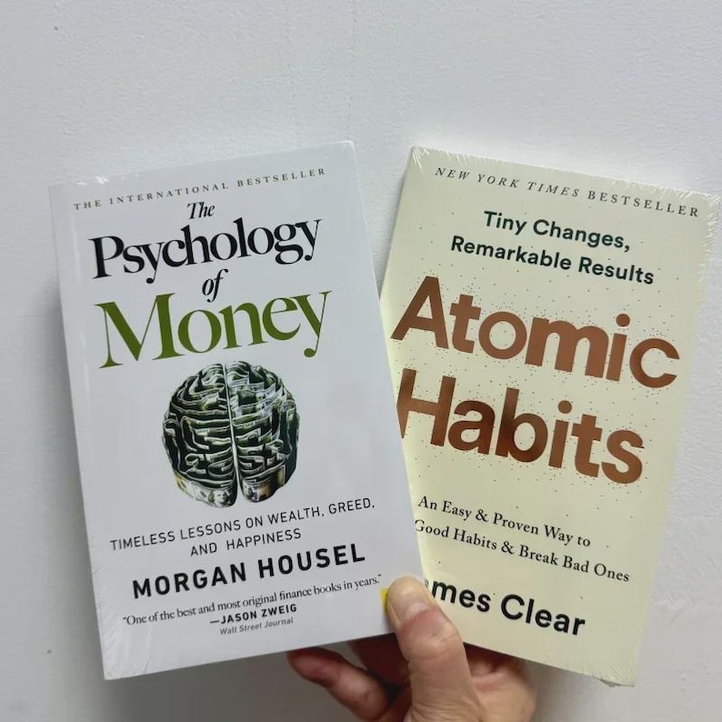 May include: Two books are displayed. "The Psychology of Money" by Morgan Housel, with a brain graphic, and "Atomic Habits" by James Clear. The books are white with black, green, and brown text. The books are on a light background.