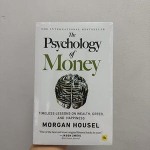 May include: A paperback book titled "The Psychology of Money" with a white cover. The title is in large green letters, with a brain graphic. The author's name is Morgan Housel. The book is described as containing timeless lessons on wealth, greed, and happiness.