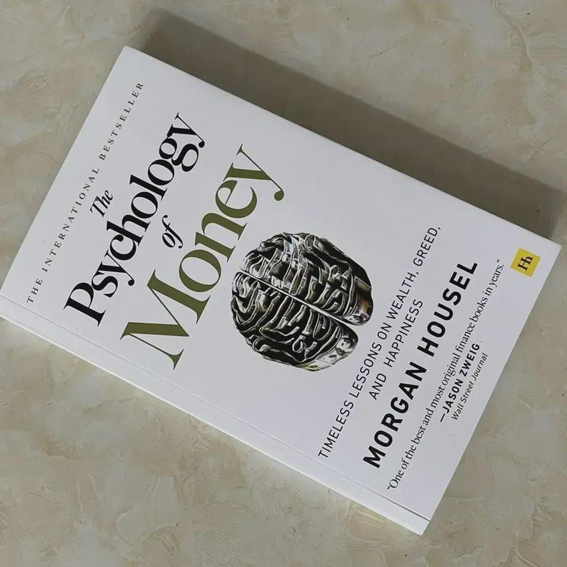 May include: A hardcover book titled "The Psychology of Money" by Morgan Housel. The cover is white with the title in large, dark green letters. A detailed, metallic-looking brain graphic is centered on the cover. The book is an international bestseller.