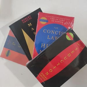 May include: A stack of hardcover books with various covers. Titles include "Mastery" and "The Concise Laws of Human Nature" by Robert Greene. The books are in different colors, including red, black, and blue, with text in white and yellow.