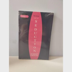 May include: A sealed paperback book titled "The Concise Art of Seduction" by Robert Greene. The book has a black cover with a vertical pink stripe down the center, displaying the title in white. An "International Bestseller" sticker is in the upper left corner.