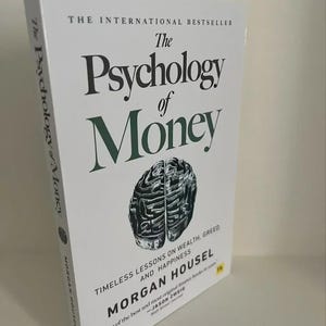 May include: A hardcover book titled "The Psychology of Money" with a white cover. The title is in large black letters with the word "Money" in green. The book features a brain graphic and author's name.