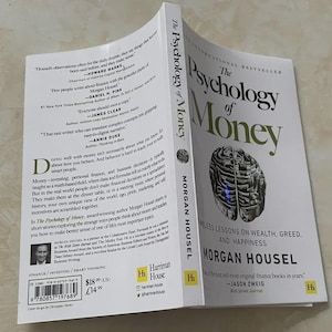 May include: A hardcover book titled "The Psychology of Money" by Morgan Housel. The cover features a stylized brain graphic and the book's title in large, bold letters. The book is open, revealing text and quotes.