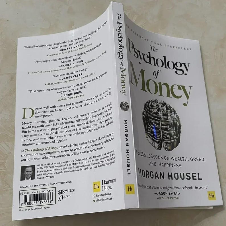 May include: A hardcover book titled "The Psychology of Money" by Morgan Housel. The cover features a stylized brain graphic and the book's title in large, bold letters. The book is open, revealing text and quotes.