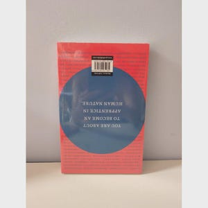 May include: A book titled "You Are About to Become an Apprentice in Human Nature" with a red cover and a blue circular design. The book has a barcode sticker on the top. The book is on a white surface.