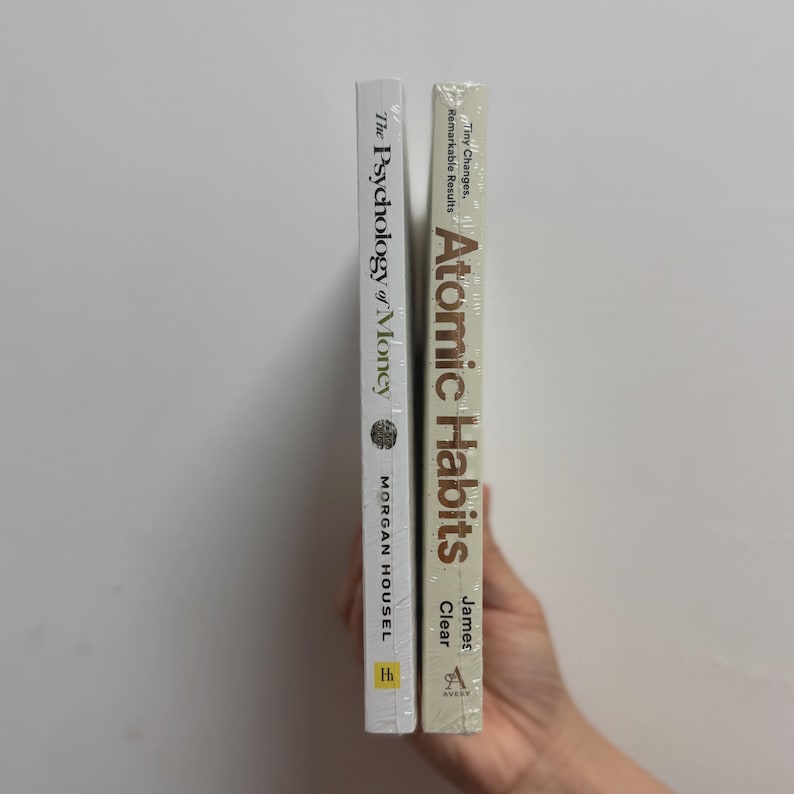 May include: Two hardcover books, "The Psychology of Money" by Morgan Housel and "Atomic Habits" by James Clear. The books are wrapped in clear plastic. The spine of "Atomic Habits" has the text "Tiny Changes, Remarkable Results". The spine of "The Psychology of Money" has the author's name.