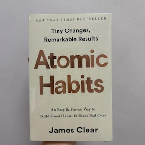 May include: A hardcover book titled "Atomic Habits" by James Clear. The cover is off-white with the title in large, copper-colored letters. The book is a New York Times bestseller and promises an easy way to build good habits and break bad ones.