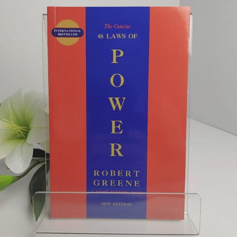 May include: A book titled "The Concise 48 Laws of Power" by Robert Greene. The book has a red cover with a blue vertical stripe down the center. The word "POWER" is printed in large white letters. An "International Bestseller" sticker is on the top left.
