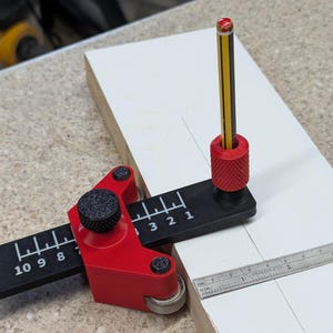 May include: A red and black woodworking marking gauge with a pencil, used for measuring and marking wood. The gauge has a black ruler with white markings and a red adjustment knob. A metal ruler is also visible.