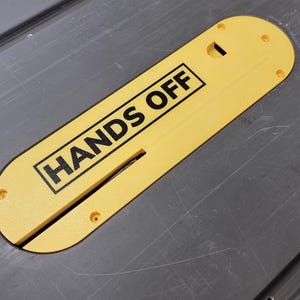 Puede incluir: Un protector de hoja de sierra de mesa amarillo con las palabras "HANDS OFF" en negro, colocado sobre una superficie de mesa gris. El protector tiene una ranura para la hoja y varios orificios para tornillos.