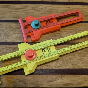 May include: Two plastic woodworking gauges in the image. One is yellow, the other is orange. Both have a ruler with measurements in millimeters, a sliding mechanism, and a circular adjustment knob. The gauges are on a wooden surface.