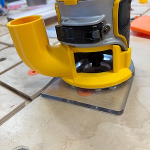May include: A yellow and gray DeWalt router with a clear plastic base is shown. The router has a yellow dust collection port and a black handle. The DeWalt logo is visible in black and yellow on the top of the tool. The router is on a wooden surface.