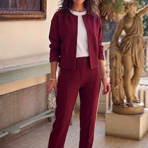 May include: A woman's burgundy suit with a matching jacket and pants. The pants have a white stripe down the side. The woman is wearing a white top, a silver clutch, and beige heels with a crystal embellishment. The background includes a painting and a statue.