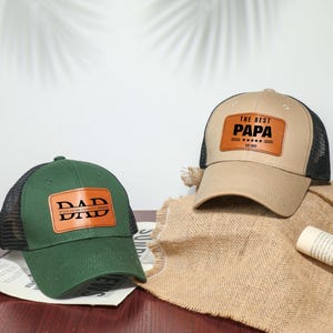 Può includere: Due cappellini da camionista con toppe in pelle. Uno è verde con "DAD" in testo bianco, l'altro è beige con "THE BEST PAPA" in testo bianco. Entrambi hanno schienali in rete nera e sono su uno sfondo neutro.
