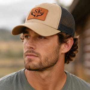 Può includere: Cappellino da camionista beige e nero con visiera curva. Presenta una toppa in pelle marrone con le iniziali "SK" e un design a corna di cervo. Il retro è in rete nera.
