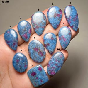 Op de afbeelding: Elf gepolijste cabochon edelstenen gelabeld A 170, met een blauwe basis met ingebedde magenta-roze insluitingen. De stenen zijn gevormd als druppels, ovalen en één vierkant, en vertonen een glad, reflecterend oppervlak met natuurlijke patronen.