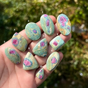 May include: Ten polished cabochon gemstones featuring a green and tan matrix with vibrant magenta-pink inclusions. Each stone is numbered from one to ten, displaying unique organic patterns and smooth, rounded surfaces in various shapes.