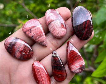 May include: Seven polished cabochon gemstones in various shapes, including oval and teardrop, featuring deep red, maroon, and pink banded patterns. Each stone is numbered from one to seven, displaying smooth, glossy surfaces with natural mineral striations.