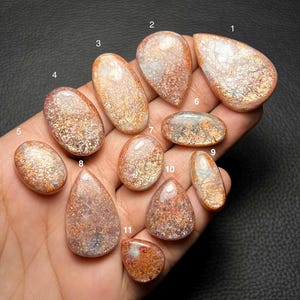 Pode incluir: Onze cabochões de pedra-do-sol polidos em várias formas, incluindo gota, oval e redondo. Cada gema exibe uma cintilação metálica de cor cobre sobre uma base translúcida de cor pêssego e branca, rotuladas com os números de 1 a 11.