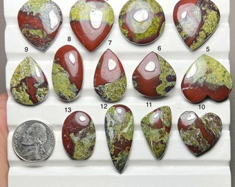 May include: A collection of thirteen polished cabochon gemstones featuring a mottled pattern of deep red and lime green. The stones are arranged on a white display tray, numbered one through thirteen, with a US nickel coin for scale.