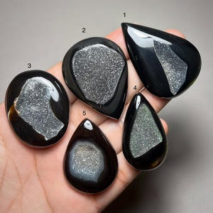 May include: Five polished black stones with sparkling, textured druzy centers, each labeled with a number from one to five. The stones vary in shape, including teardrop and oval forms, and feature smooth, glossy surfaces surrounding the crystals.