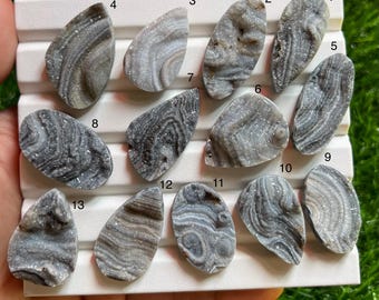 Può includere: Tredici cabochon in agata grigia e bianca con texture di cristalli druzy, disposti su un vassoio espositore bianco. Ogni pietra presenta motivi a bande unici ed è numerata da uno a tredici, mostrando forme varie tra cui goccia e ovale.