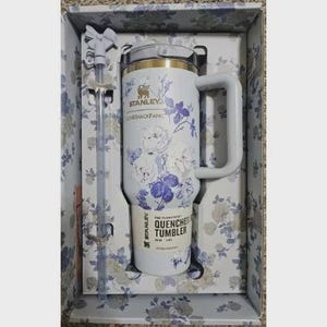 Op de afbeelding: Een witte Stanley Quencher-beker met een bloemenontwerp in een geschenkdoos. De beker heeft een handvat en een rietje. Het bloemmotief omvat witte en paarse bloemen. De doos heeft een bijpassend bloemmotief.