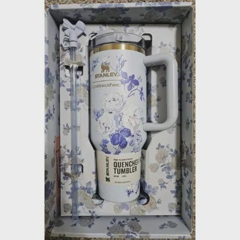 Op de afbeelding: Een witte Stanley Quencher-beker met een bloemenontwerp in een geschenkdoos. De beker heeft een handvat en een rietje. Het bloemmotief omvat witte en paarse bloemen. De doos heeft een bijpassend bloemmotief.