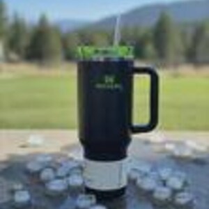 May include: A black insulated travel mug with a green lid and a white straw. The mug has a black handle and a white base. The mug is sitting on a table with ice cubes. The mug has a logo on the side.