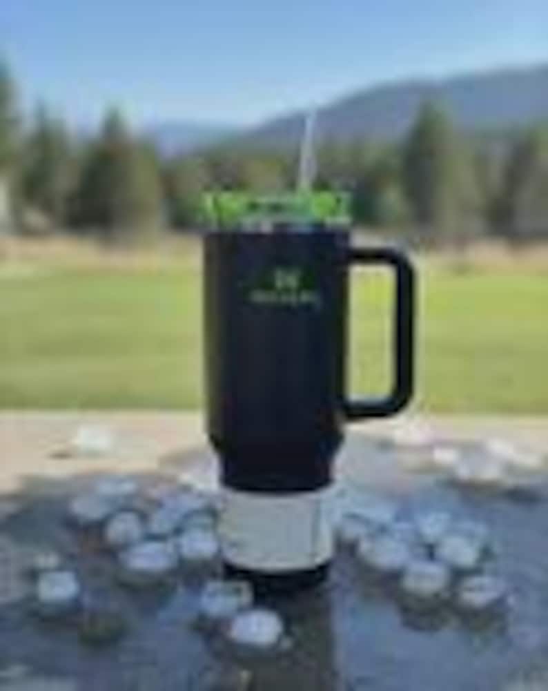 May include: A black insulated travel mug with a green lid and a white straw. The mug has a black handle and a white base. The mug is sitting on a table with ice cubes. The mug has a logo on the side.