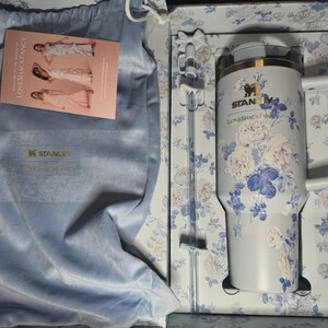 Op de afbeelding: Een Stanley Loveshackfancy tumbler met een witte handgreep en bloemenpatroon, gepresenteerd in een doos met een bijpassend bloemenpatroon. Inclusief een lichtblauwe fluwelen tas en een kaart met de tekst "Welcome to the world of Loveshackfancy".