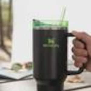 May include: A black insulated travel mug with a green lid and handle. The mug has a white base and a straw. The text "HydroJug" is printed in green on the side. The mug is held by a person.
