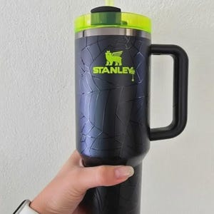 May include: Black and silver Stanley tumbler with a handle, a green lid, and a green straw. The tumbler has a cracked pattern design and the Stanley logo in green. The tumbler is held by a person.