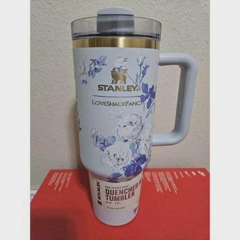 Op de afbeelding: Een lichtblauwe Stanley Quencher-beker met een bloemenpatroon in wit en blauw. De beker heeft een handvat, een doorzichtig deksel en gouden accenten. De tekst "STANLEY" en "LOVESHACKFANCY" zijn zichtbaar.