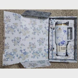 Op de afbeelding: Een witte Stanley Quencher tumbler met een bloemenontwerp in een geschenkverpakking. De tumbler heeft een handvat en een rietje. De doos en de tumbler hebben een patroon van blauwe en witte bloemen.