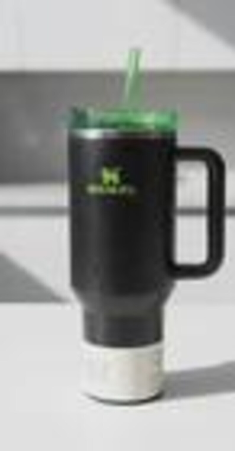 May include: A black insulated tumbler with a green lid and straw. The tumbler has a white base and a black handle. The Stanley logo is printed in green on the side. The tumbler is designed to keep drinks cold or hot.