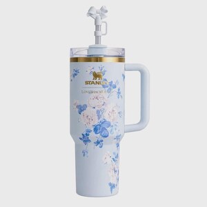 Puede incluir: Un vaso t&eacute;rmico azul claro con asa, que presenta un estampado floral blanco, el logotipo dorado de Stanley y el texto LoveShackFancy. La tapa incluye una pajita transparente rematada con un accesorio decorativo de lazo blanco.