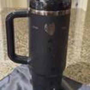 Pu&ograve; includere: Un bicchiere termico nero opaco con una robusta maniglia laterale e un coperchio in plastica trasparente. Il contenitore cilindrico presenta un emblema argentato metallizzato sulla parte anteriore superiore e una base affusolata per le bevande.