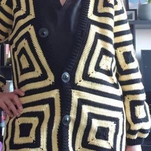 Può includere: Cardigan all'uncinetto nero e beige con motivo geometrico. Il cardigan ha uno scollo a V, maniche lunghe e grandi bottoni neri. Il design presenta quadrati dentro quadrati, creando un look audace. Capo fatto a mano.