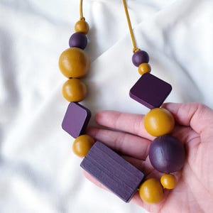 Peut inclure: Un collier composé de perles en bois de couleurs moutarde et violet. Le collier est composé de perles rondes et carrées enfilées sur un cordon beige. Le collier est tenu dans une main sur fond blanc.