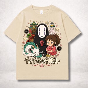 Op de afbeelding: Beige T-shirt met een kleurrijke afbeelding uit de animatiefilm Spirited Away. Het ontwerp toont personages als No-Face, Chihiro en Haku, samen met Japanse tekst. Het shirt heeft een ronde hals en korte mouwen.