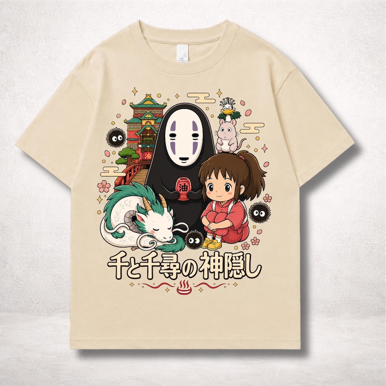 Op de afbeelding: Beige T-shirt met een kleurrijke afbeelding uit de animatiefilm Spirited Away. Het ontwerp toont personages als No-Face, Chihiro en Haku, samen met Japanse tekst. Het shirt heeft een ronde hals en korte mouwen.